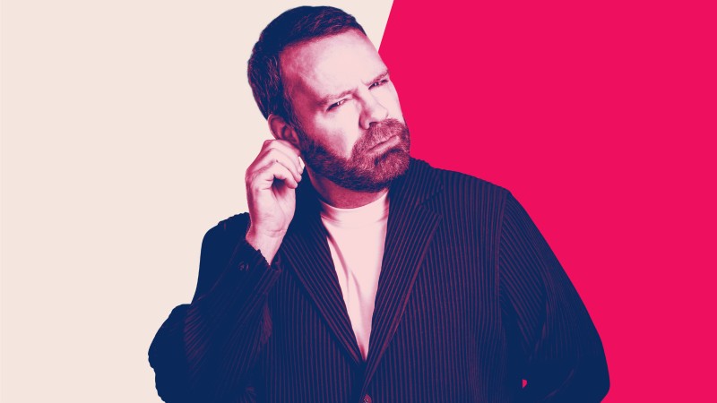 Neil Delamere against a pink and black backdrop. He is scratching his ear and looks like he is thinking.