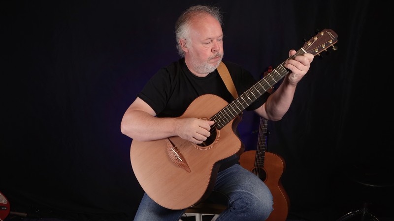 Paul Rose, sitting playing an acoustic guitar against a black background.
