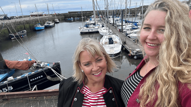 The Salty Scrimshaws syand in the forground smiling at the camera with a marina as a backdrop.