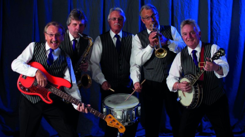 The five members of Clubsound, suited and booted, wielding their instruments, smiles on their faces, posing for a promo photo in front of a blue backdrop