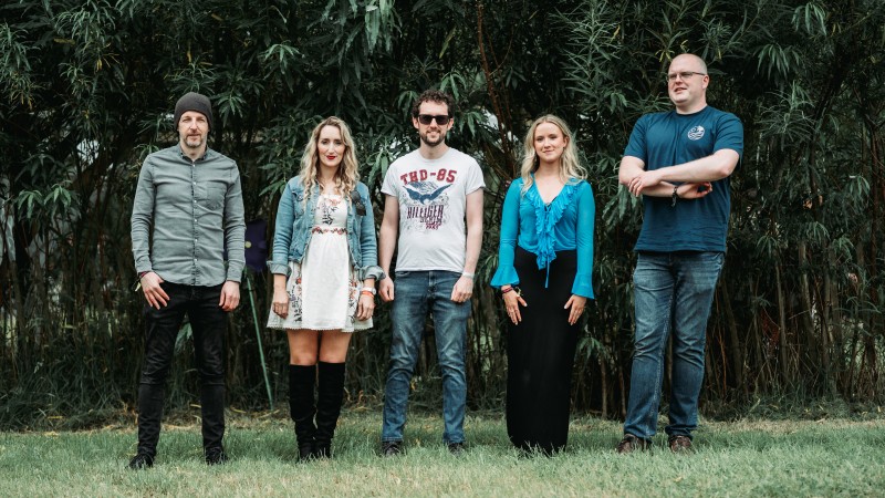 All five members of Lore standing smiling at the camera, standing on short grass in front of a backdrop of dense woodland.