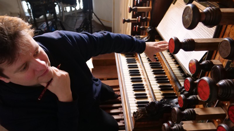 Daniel Moult, wearing a blue suit studying the controls of a pipe organ while sat at the keyboard
