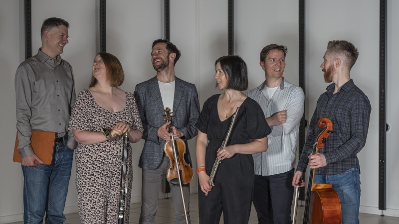 The six members of the Hard Rain Ensembble, 4 men and 2 women, standing talking and laughing while each is holding their instrument, standing in front of a white wall will black beams.