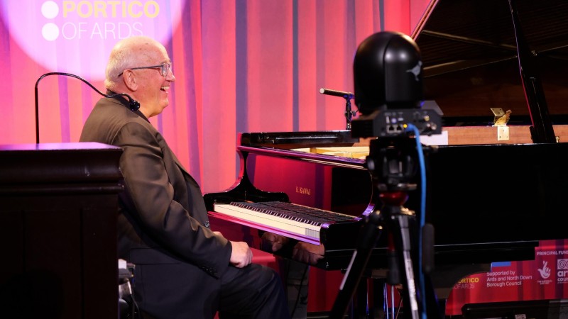 John, wearing a brown jacket and glasses photographed candidly at a grand or baby grand piano in Portico or Ards preparing to play.