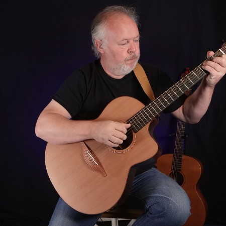 Paul Rose, sitting playing an acoustic guitar against a black background.
